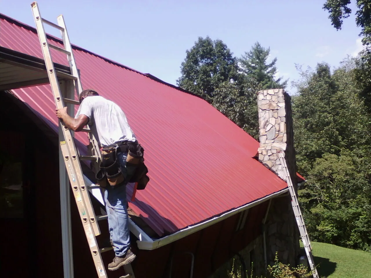 Roofer on a ladder beside a red metal roof and stone chimney — Soffit & Fascia Repair by Roofing Brothers in Orrville